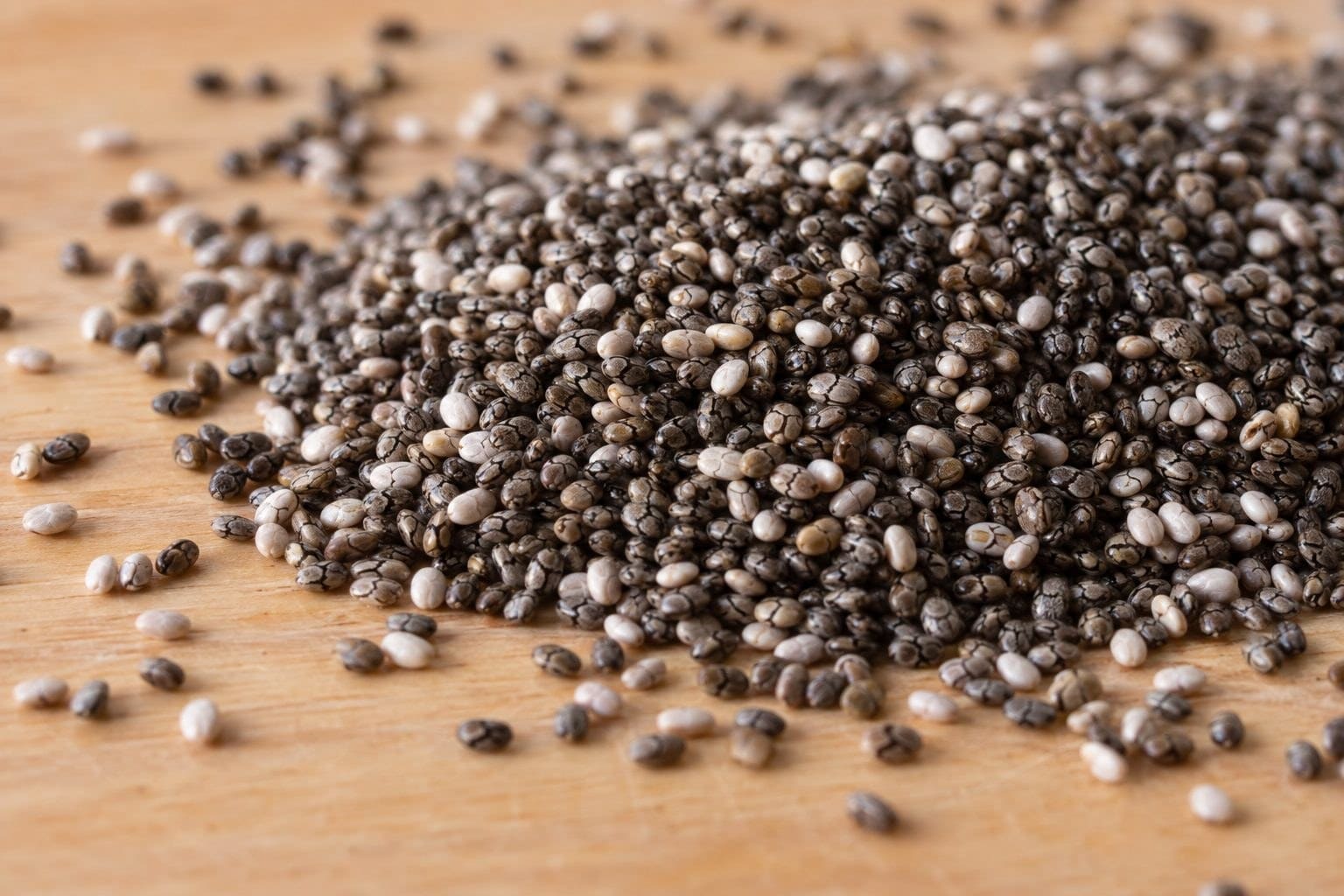 Graines de chia noires et blanches en gros plan, dispers&eacute;es sur une surface naturelle en bois clair