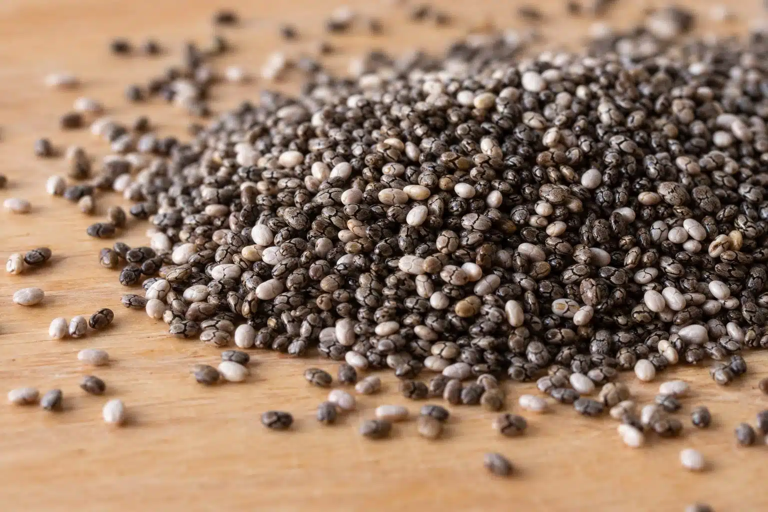Graines de chia noires et blanches en gros plan, dispers&eacute;es sur une surface naturelle en bois clair